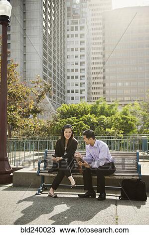 Stock Image - Chinese business people using technology on city bench. Fotosearch