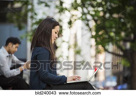 Stock Image - Chinese businesswoman typing on laptop in city. Fotosearch