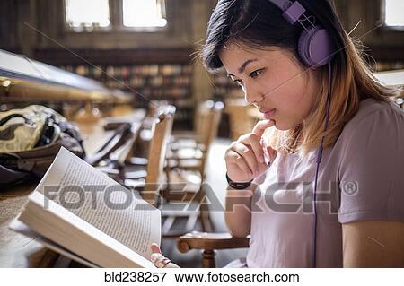 Stock Photo - Chinese woman in library reading book and listening to headphones. Fotosearch