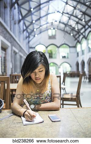 Stock Photograph - Chinese woman sitting in library writing in notebook. Fotosearch