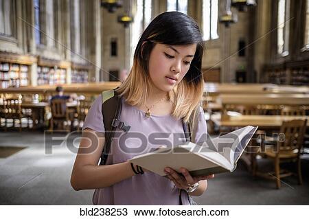 Stock Image - Chinese woman standing in library reading book. Fotosearch