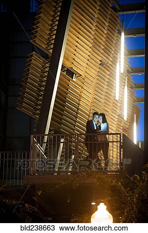 Stock Image - Couple standing on balcony posing for cell phone selfie at night. Fotosearch