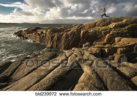Stock Image - Distant Caucasian woman doing yoga on rocks near ocean. Fotosearch