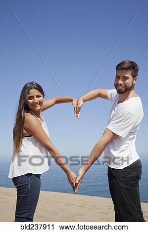 Stock Image - Hispanic couple making heart-shape with arms at beach. Fotosearch