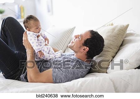 Stock Photo - Hispanic father playing with baby daughter on bed. Fotosearch