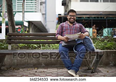 Stock Image - Hispanic man sitting on bench reading brochure. Fotosearch