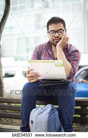 Stock Image - Hispanic man sitting on top of bench reading brochure. Fotosearch