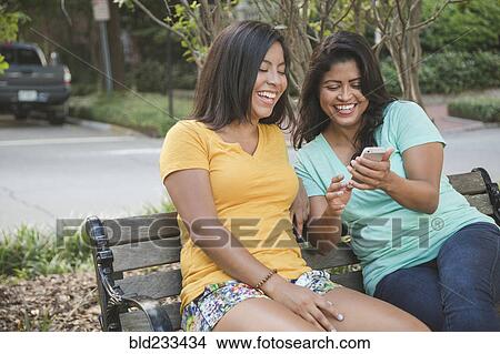Picture - Hispanic mother and daughter using cell phone on park bench. Fotosearch