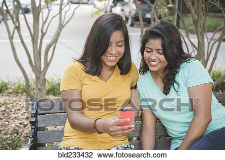 Stock Image - Hispanic mother and daughter using cell phone on park bench. Fotosearch