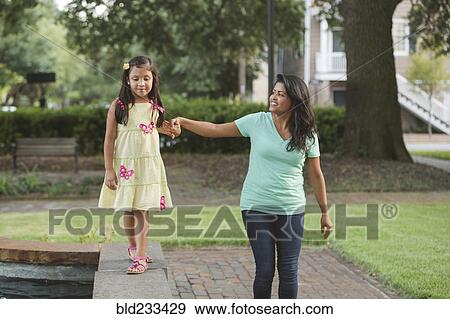 Stock Photo - Hispanic mother holding hand of daughter walking on park wall. Fotosearch