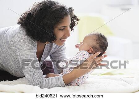 Stock Image - Hispanic mother laying on bed holding baby daughter. Fotosearch
