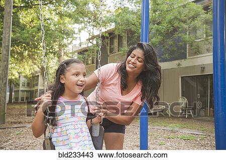 Stock Image - Hispanic mother pushing daughter in playground swing. Fotosearch