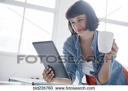 Stock Image - Hispanic woman drinking coffee and reading digital tablet. Fotosearch