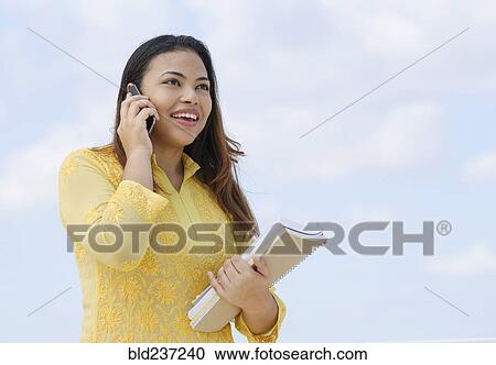Stock Image - Hispanic woman holding notebooks talking on cell phone. Fotosearch