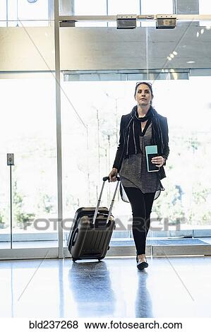 Stock Photo - Hispanic woman holding passport pulling suitcase in airport. Fotosearch