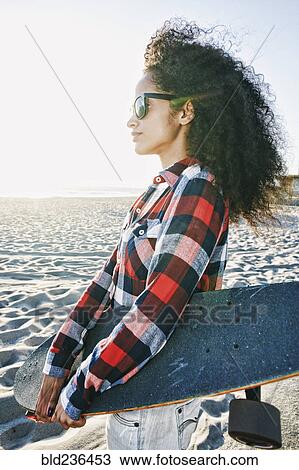 Stock Image - Hispanic woman holding skateboard at beach. Fotosearch