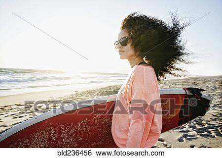 Stock Photograph - Hispanic woman holding surfboard at beach. Fotosearch