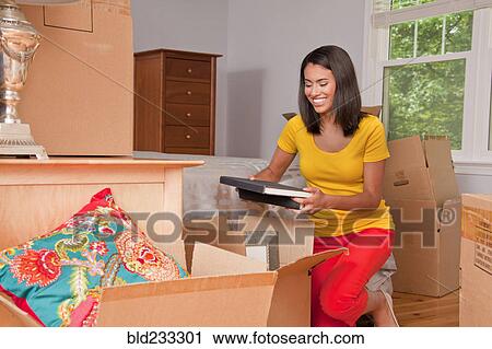 Stock Image - Hispanic woman kneeling on floor packing books in moving boxes. Fotosearch