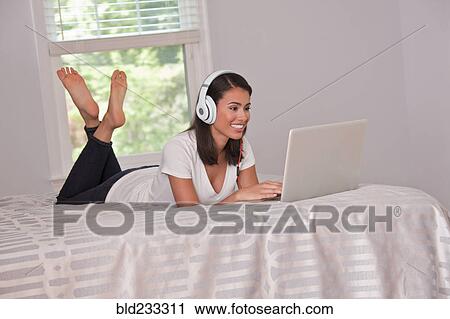 Hispanic woman laying on bed listening to laptop with headphones View Large Photo Image Stock Image - Hispanic woman laying on bed listening to laptop with headphones. Fotosearch