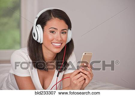 Hispanic woman laying on bed listening to cell phone with headphones View Large Photo Image Stock Photo - Hispanic woman laying on bed listening to cell phone with headphones. Fotosearch