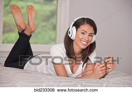 Stock Photo - Hispanic woman laying on bed listening to cell phone with headphones. Fotosearch