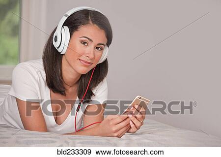 Stock Photo - Hispanic woman laying on bed listening to cell phone with headphones. Fotosearch