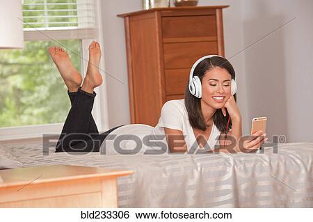 Stock Photograph - Hispanic woman laying on bed listening to cell phone with headphones. Fotosearch