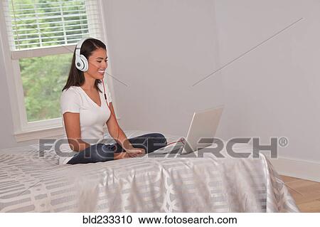 Stock Image - Hispanic woman sitting on bed listening to laptop with headphones. Fotosearch