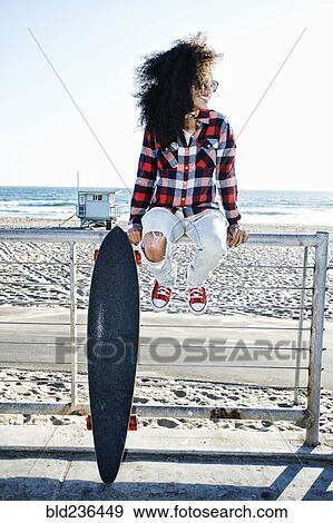 Stock Photo - Hispanic woman sitting on fence at beach with skateboard. Fotosearch