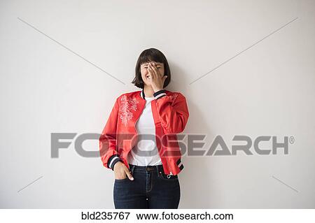 Stock Photo - Laughing Hispanic woman wearing red jacket leaning on wall. Fotosearch