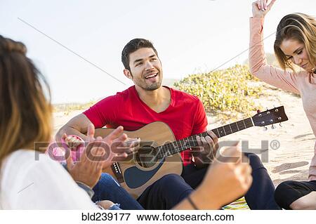 Stock Photo - Man playing guitar for friends at beach. Fotosearch