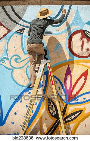 Stock Photograph - Man standing on ladder painting mural on wall. Fotosearch