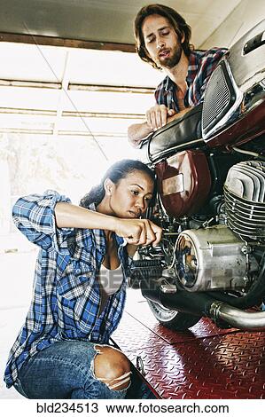 Stock Image - Man watching woman repairing motorcycle in garage. Fotosearch