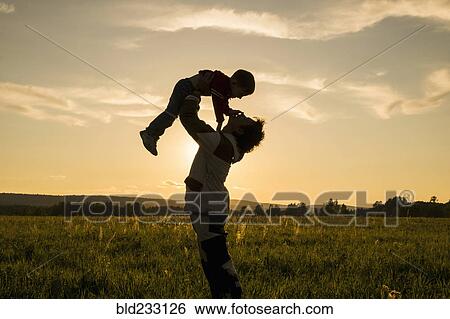 Stock Photograph - Mari man lifting son in field at sunset. Fotosearch