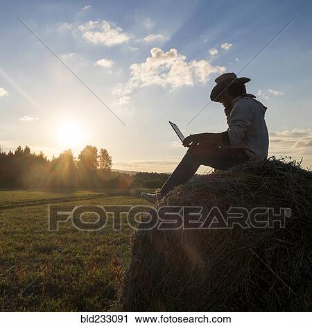 Stock Image - Mari man sitting on hay bale at sunset using laptop. Fotosearch