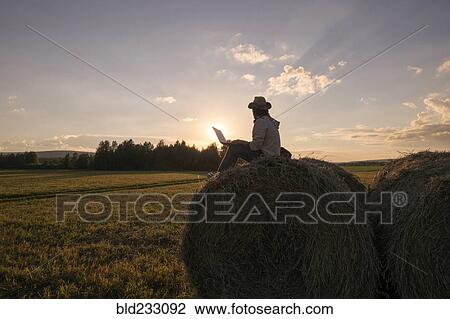Stock Image - Mari man sitting on hay bale at sunset using laptop. Fotosearch