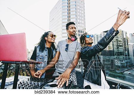 Picture - Men posing for cell phone selfie with DJ on urban rooftop. Fotosearch