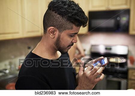 Stock Image - Middle Eastern man texting on cell phone in kitchen. Fotosearch