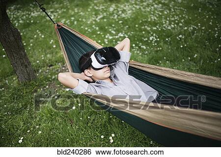 Stock Photograph - Mixed Race boy laying in hammock wearing virtual reality goggles. Fotosearch