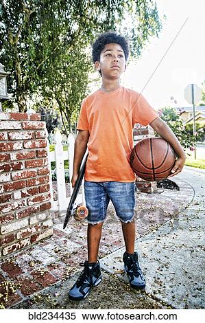 Stock Photography - Mixed Race boy standing on sidewalk holding skateboard and basketball. Fotosearch