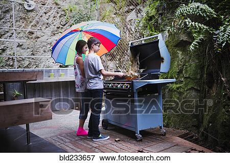 Stock Photo - Mixed Race brother and sister cooking on patio grille under umbrella. Fotosearch
