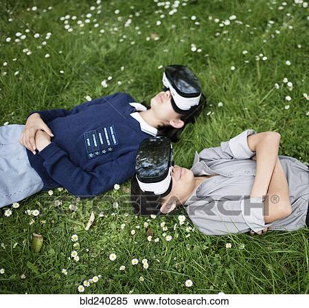 Stock Photography - Mixed Race brother and sister laying in grass wearing virtual reality goggles. Fotosearch