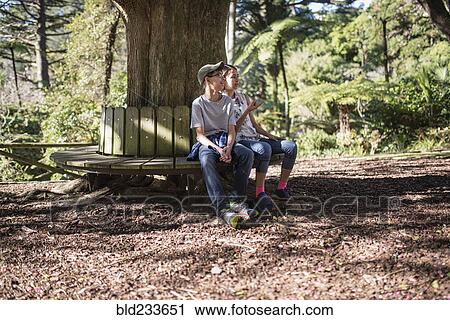 Stock Image - Mixed Race brother and sister sitting on bench at tree. Fotosearch