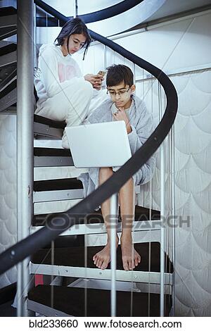 Stock Image - Mixed Race brother and sister sitting on spiral staircase using laptops. Fotosearch