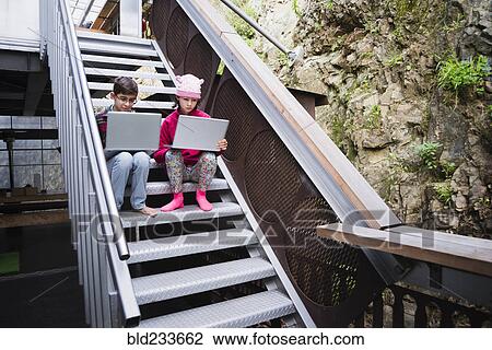 Stock Image - Mixed Race brother and sister sitting on staircase using laptops. Fotosearch