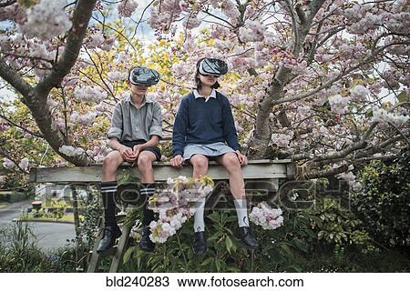 Stock Image - Mixed Race brother and sister sitting in tree wearing virtual reality goggles. Fotosearch
