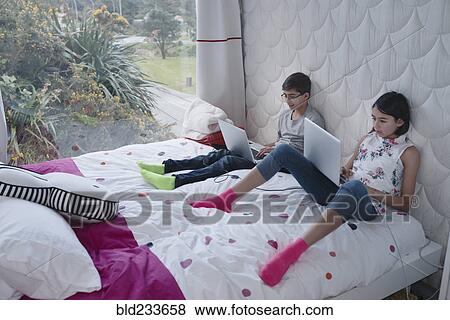 Stock Photo - Mixed Race brother and sister sitting on bed using laptop. Fotosearch
