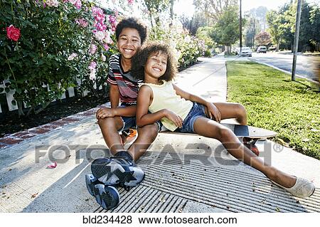 Stock Photo - Mixed Race brother and sister sitting on skateboard on sidewalk. Fotosearch