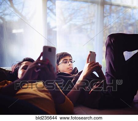 Picture - Mixed Race brother and sister texting on cell phones near window. Fotosearch