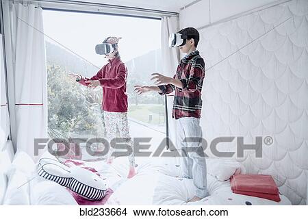 Picture - Mixed Race brother and sister using virtual reality goggles on bed. Fotosearch